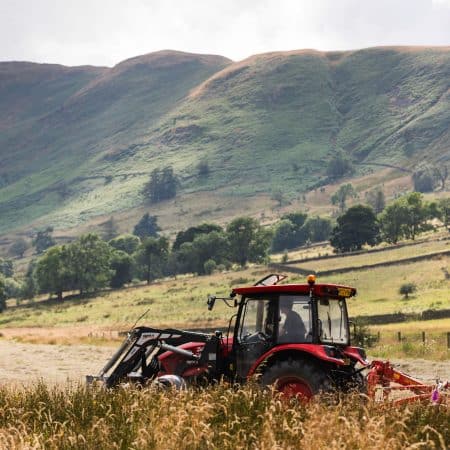 Tractor in a hay meadow pulling a hay tedder in mountainous surroundings.